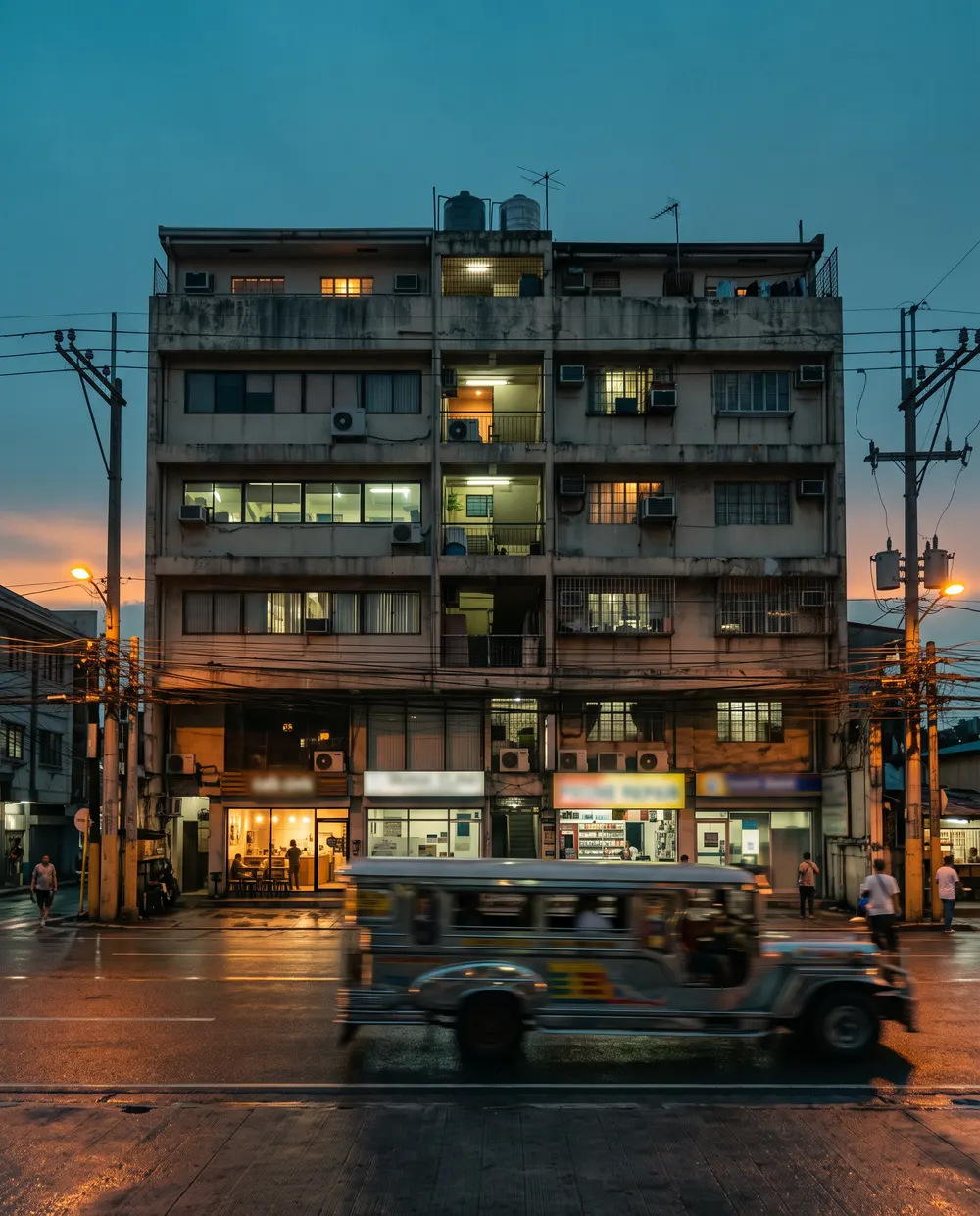 A six-storey mixed-use building with lit ground-floor tenants at dusk